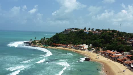 Dolly-out-rising-aerial-drone-shot-of-the-tropical-famous-Baia-Formosa-beach-town-in-the-state-of-Rio-Grande-do-Norte,-Brazil-with-fishing-boats,-coastal-homes,-small-waves,-and-surfers