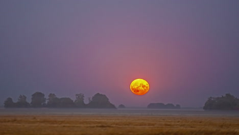 super moon rises over a farm field with a low layer of fog or mist to glow over the countryside - time lapse