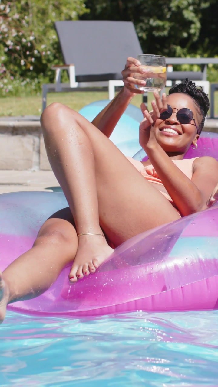 Vertical video: Relaxing woman in bikini sipping on float at home pool man entering giving glass
