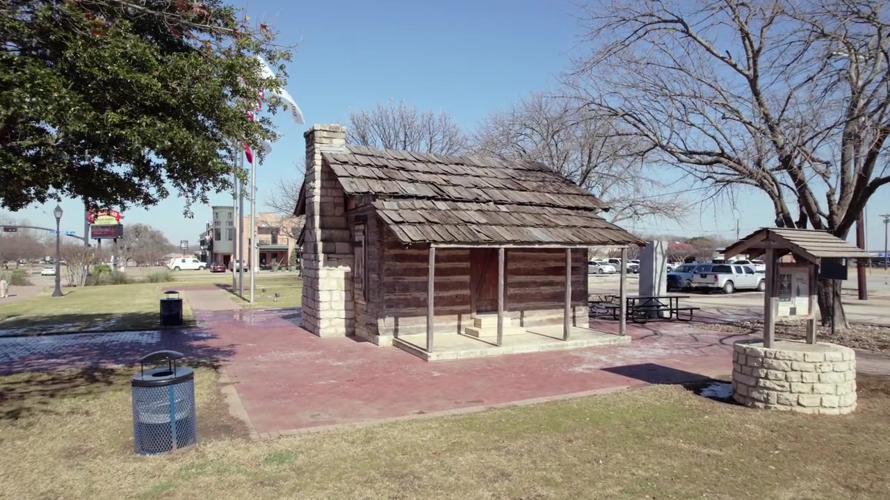 Filmagem 4K de drone com aproximação da histórica Cabana de Toras Larkin Newton no centro de Midlothian, Texas, com bandeiras americana e de Midlothian tremulando ao vento ao lado do marco.
