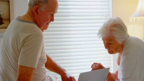 vista lateral de una pareja de ancianos caucásicos usando una computadora portátil en la cama en el dormitorio en una cómoda casa de 4k