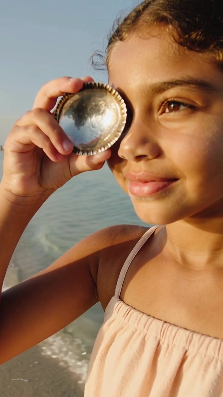 Video verticale: Bambina in vestito rosa che inclina uno specchio di conchiglia verso l'occhio destro in spiaggia