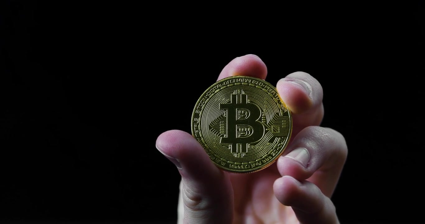 Uma mão segura um bitcoin dourado brilhante em foco nítido contra um fundo preto dramático