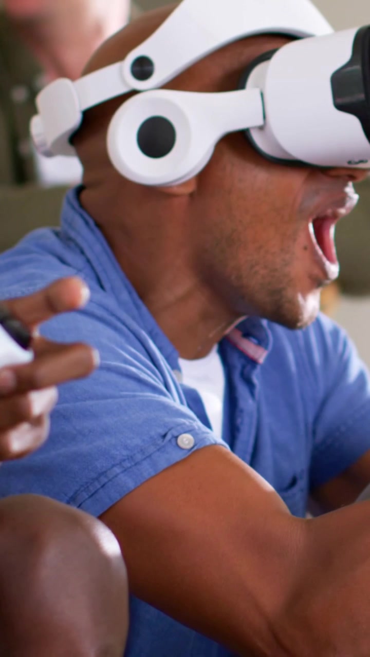 Vertical video: Putting on white VR set, blue-shirt man reacting at home holding gamepad to play