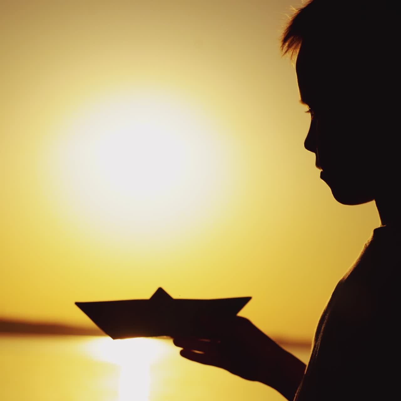 Silhouette of a little boy is holding a paper boat in his hand and looking at it on the background of sunset in the summer near the pond. Close-up