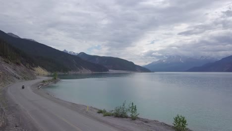 Un-Viajero-Motociclista-Conduciendo-Por-Una-Carretera-Con-Curvas-Al-Lado-De-Un-Impresionante-Lago-Canadiense-Con-Una-Cordillera-A-Los-Lados-En-Un-Día-Nublado,-Tiro-Panorámico-Bloqueado