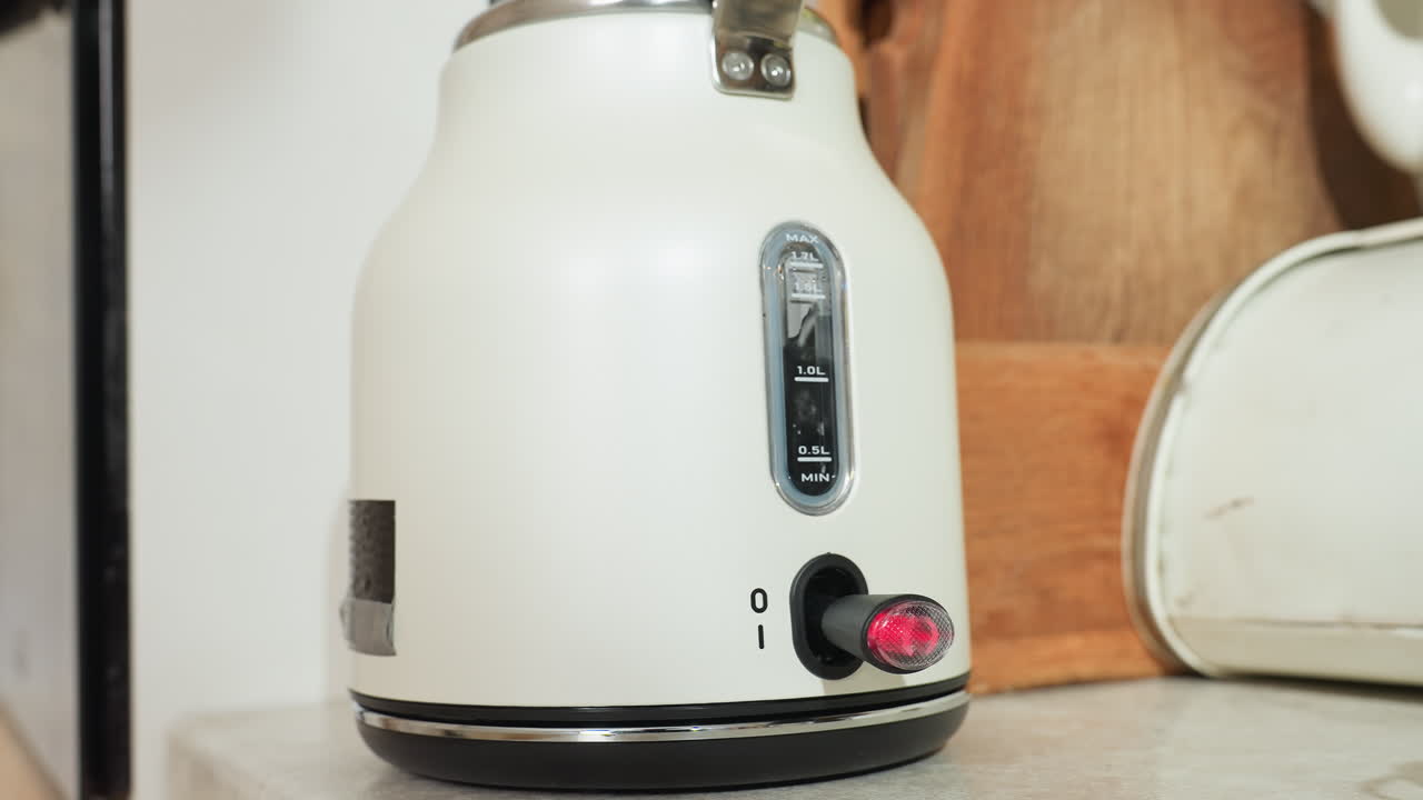 Close up of electric kettle with visible water level steaming inside and power indicator blinking red while sitting on countertop beside wooden spatula and vintage bread box in softly lit kitchen