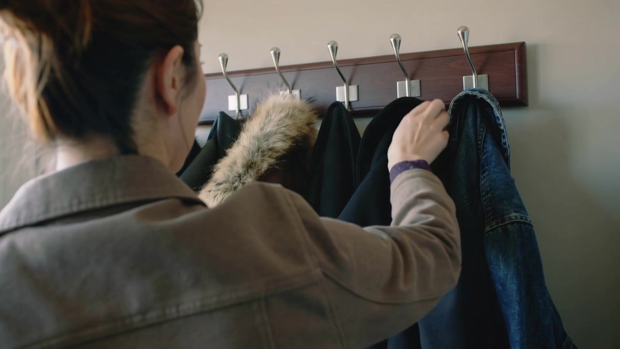 Una donna che entra, in trench color cammello, allunga la mano verso un cappotto scuro su uno stender in legno, facendo spazio.