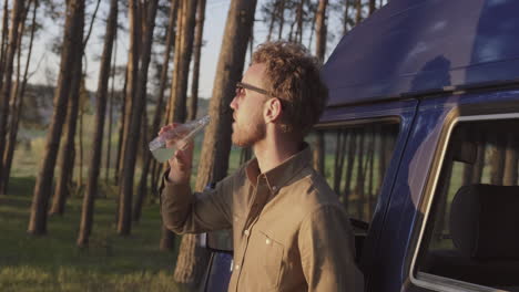 a relaxed guy takes a drink next to a caravan in the middle of the forest