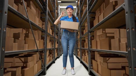 full body of asian female courier in blue uniform showing okay gesture and smiling while delivering a carton in warehouse