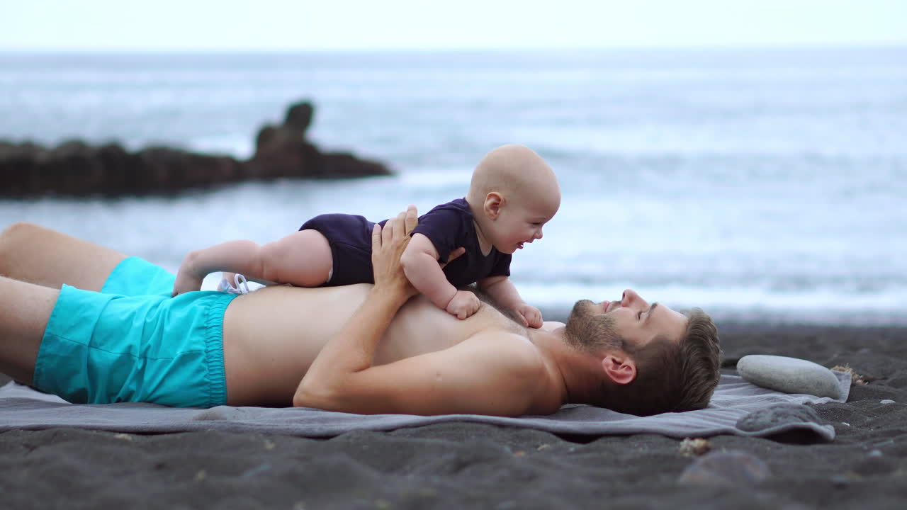 Amidst the beach setting, a young father delights in playful bonding with his newborn son. These special moments enhance his vacation experience. He embodies the joy of fatherhood