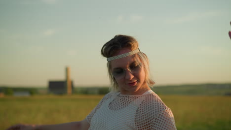 a joyful woman wearing a headband and sunglasses dances freely, raising her hands in an open grassy field, while the blurred figure of someone else can be seen in the background