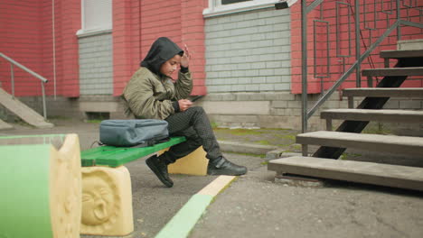 Boy in hooded jacket seated on green bench outdoors holding phone, adjusting hoodie cap with hand, gray backpack beside him, near brick building and outdoor steps in urban environment