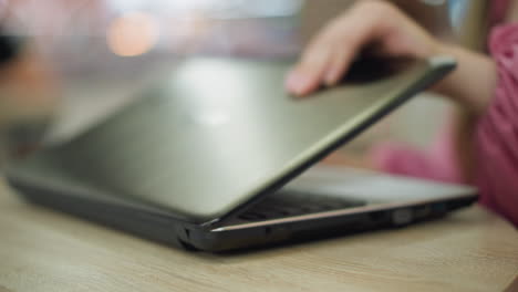 hand of lady in pink dress and wrist chain gently places laptop on wooden table and begins opening it, preparing to work, while the background is blurred, with environment when lit up