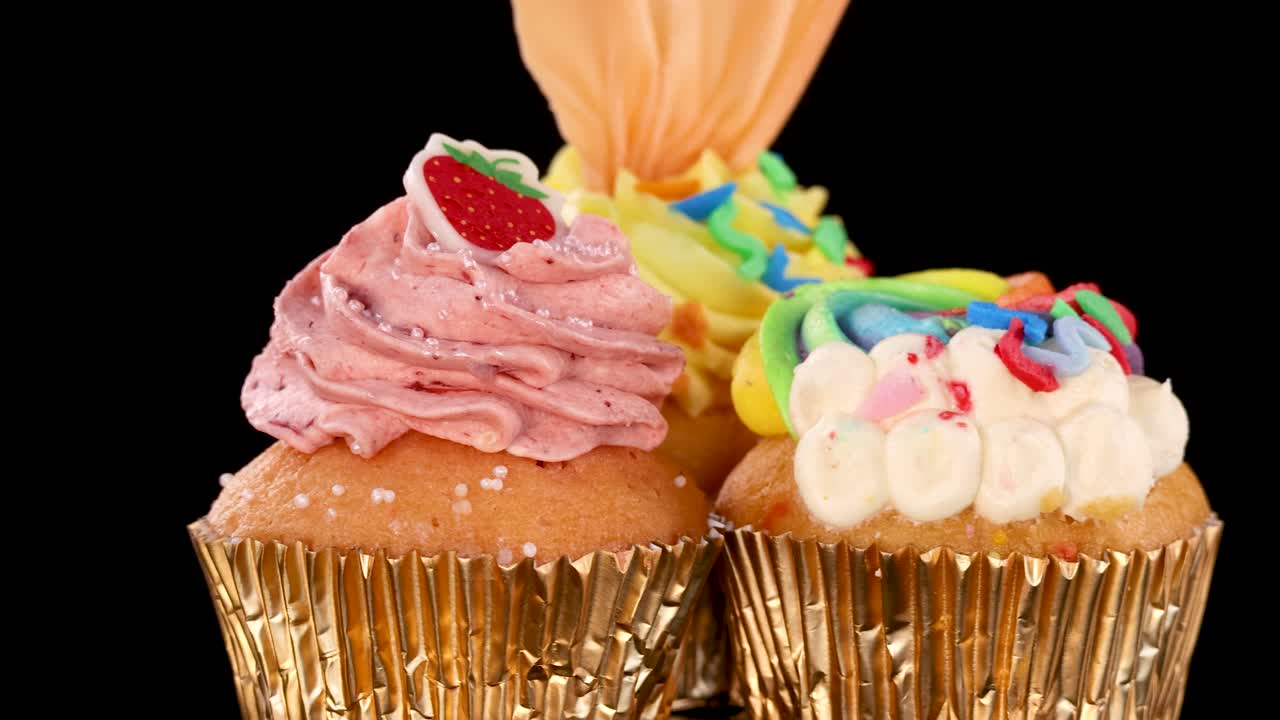 Three elaborately decorated cupcakes with rainbow and strawberry toppings rotate smoothly against a seamless black background, under bright, even studio lighting
