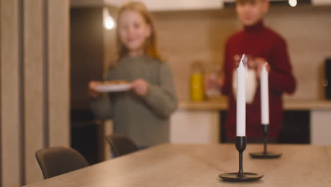 Un-Niño-Rubio-Y-Su-Hermana-Rubia-Colocan-Un-Vaso-Vacío,-Una-Jarra-De-Leche-Y-Un-Plato-Lleno-De-Galletas-En-Una-Mesa-Vacía-Con-Dos-Velas,-Luego-Salen-De-La-Habitación-3