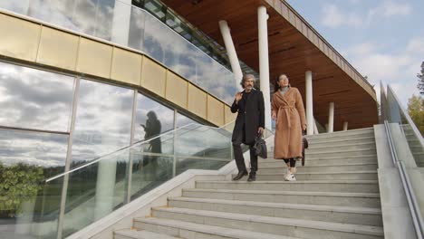 young businesswoman in elegant beige coat talking with coworker while going down the stairs