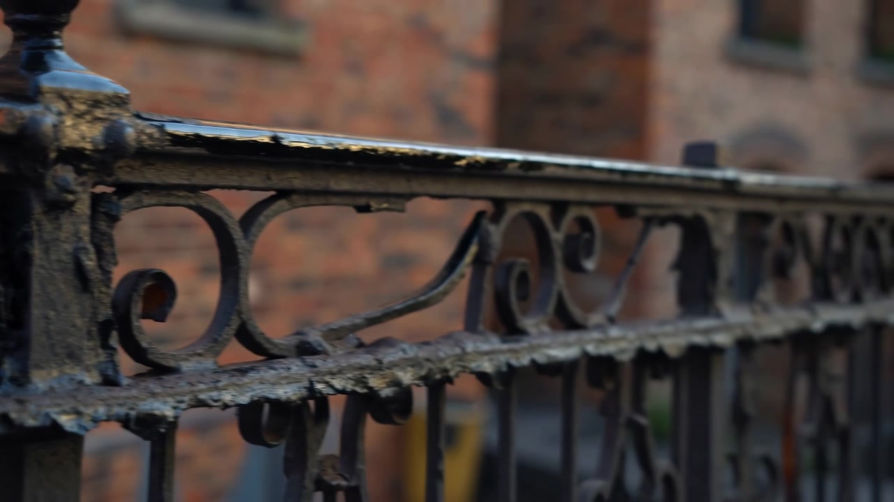Ornamentalt smedejernsgelænder i nærbillede mod blød murstenssløring, der viser afskallende maling, afskallende kanter, rustpletter, snavs og gradvis forvitring på tværs af metaloverfladen og snirklerne