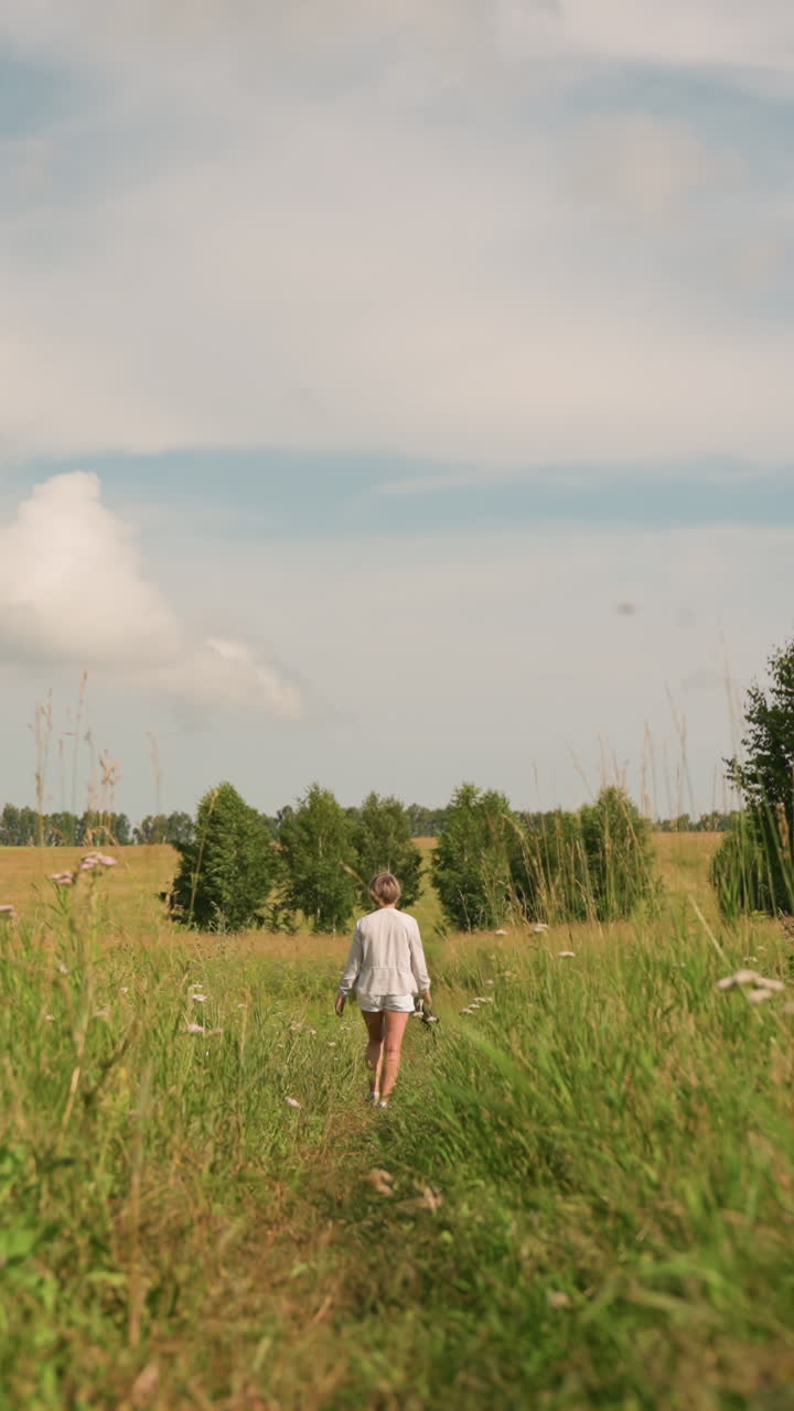 하 옷을 입은 짧은 여자가 그녀의 손에 무언가를 들고 높은 잔디을 걸어가는 장면은 산란한 구름과 함께 밝은 하늘 아래에 설정되어 있으며, 녹색 식물과 꽃으로 둘러싸여 있습니다.