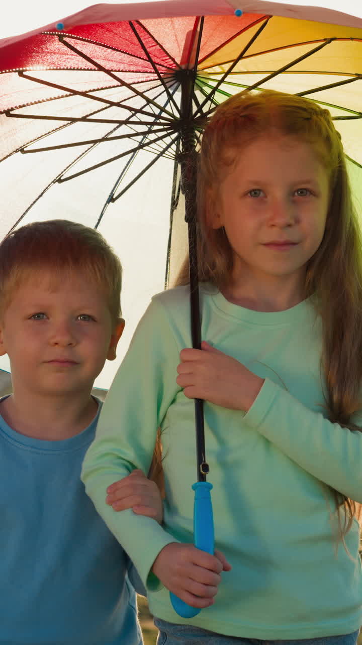 Children under umbrella at riverside on rainy day. Cute little brother and sister hide together under rainbow parasol from raindrops. Boy and girl in nature