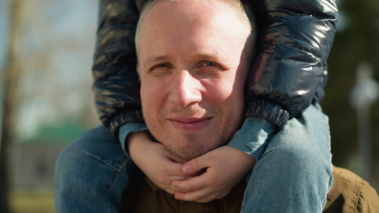 un hombre sonriendo mientras lleva a un niño en sus hombros, el hombre está vestido con una chaqueta marrón, y el niño está envuelto en una chaqueta negra, con una vista borrosa de los árboles