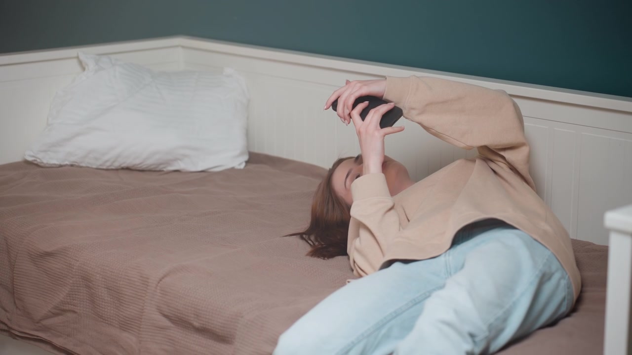 Ung person ligger på sengen og scroller på telefonen under fjernstudiepause, afslappet trøje og jeans, neutralt sengetøj og pude, afdæmpet turkis væg, distraheret fra grammatikøvelse, blødt naturligt lys, stille.
