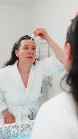 middleaged woman applying facial serum drops gently on skin smiling satisfied mirror reflection