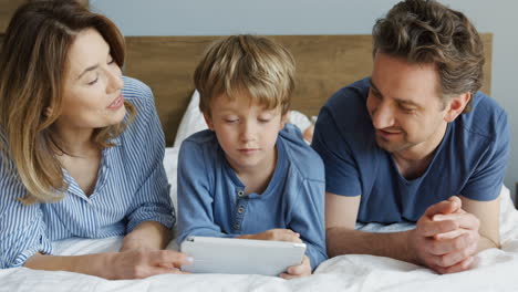 vista de cerca de un niño pequeño y sus padres acostados en la cama y viendo algo en la tableta