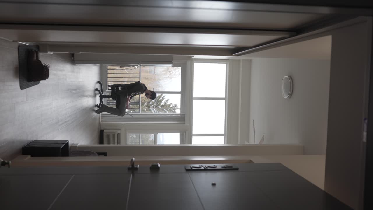 video vertical, joven con computadora portátil y auriculares sentado en una silla en una habitación luminosa, escuchando música en línea, estática, vista de puerta abierta