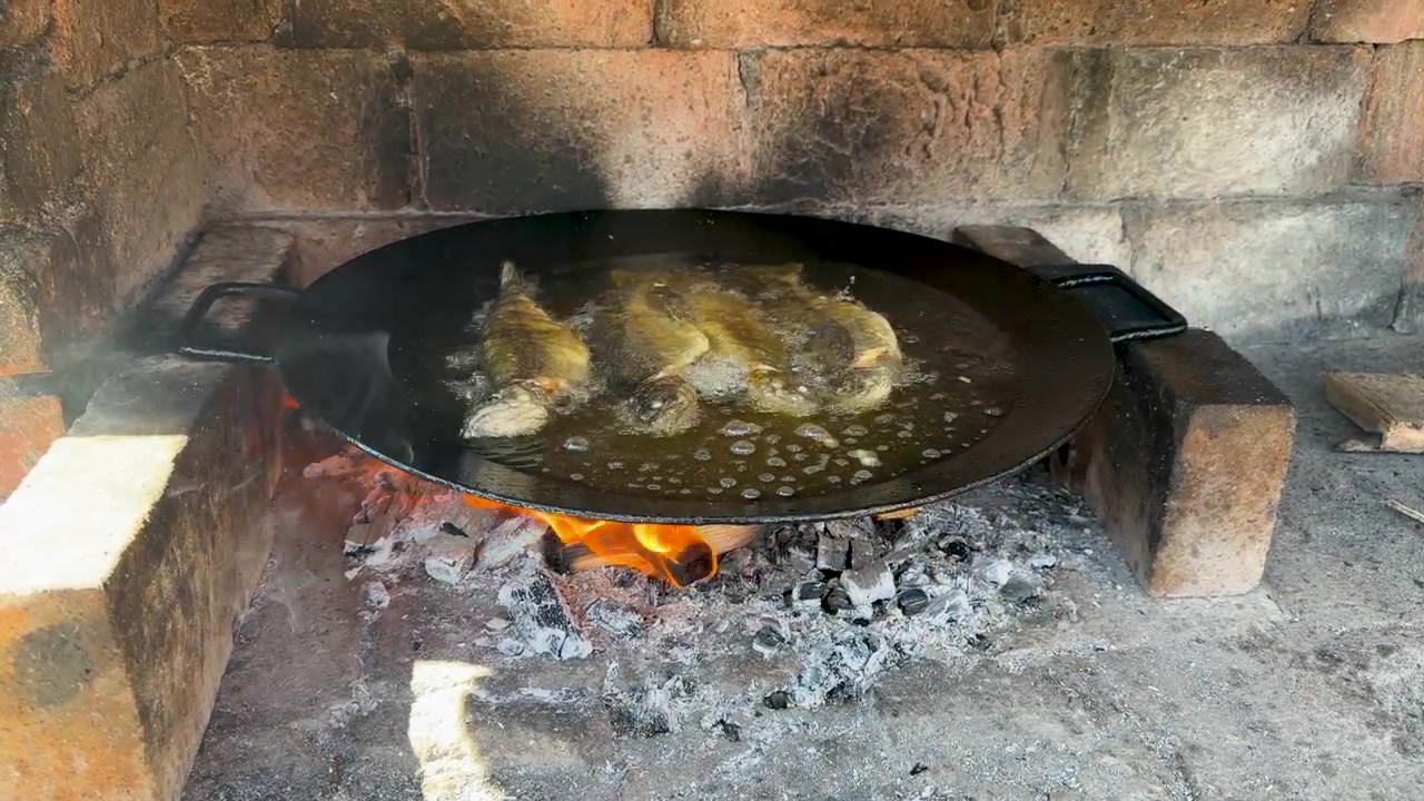 Diversi pesci interi friggono in olio bollente in un'ampia padella nera su un fuoco a legna aperto, con fiamme, braci ardenti, cenere grigia, supporti in mattoni e vapore che sale attorno al focolare rustico