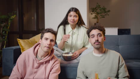 three friends watching comic movie on television sitting on couch and floor, eating popcorn and drinking soda