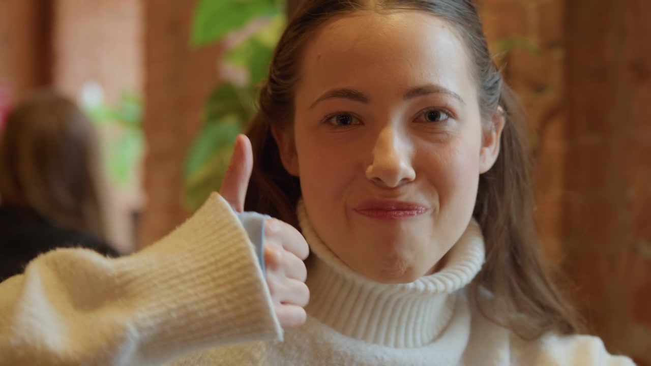 Close up of woman with long brown hair wearing white turtleneck sweater in indoor setting. subject remains mostly stationary occasionally moving her head slightly and giving thumbs