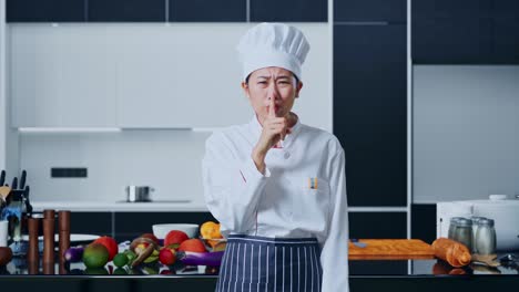 mujer chef asiática mirando a la cámara y haciendo un gesto de silencio en la cocina de casa