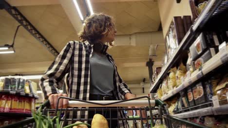 Smiling-Woman-In-Plaid-Shirt-And-Headphones-On-Neck-Is-Walking-In-Grocery-Store-Steering-Shopping-Trolley-With-Food-Inside