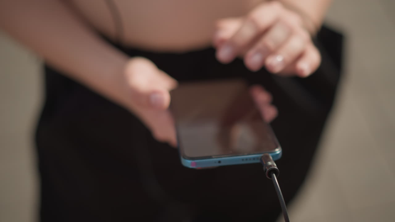 Giovane donna caucasica che tiene in mano uno smartphone su un marciapiede soleggiato, districando il cavo degli auricolari e infilando il telefono in tasca tra le prove di danza, indossa un top corto bianco e pantaloni neri, primo piano sulle mani
