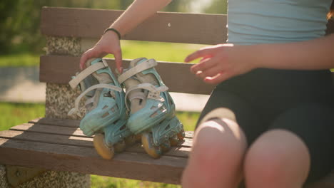 young white lady drops cyan roller skates on wooden bench and sits beside it with abdomen slightly exposed, sunlight reflects on her with greenery background