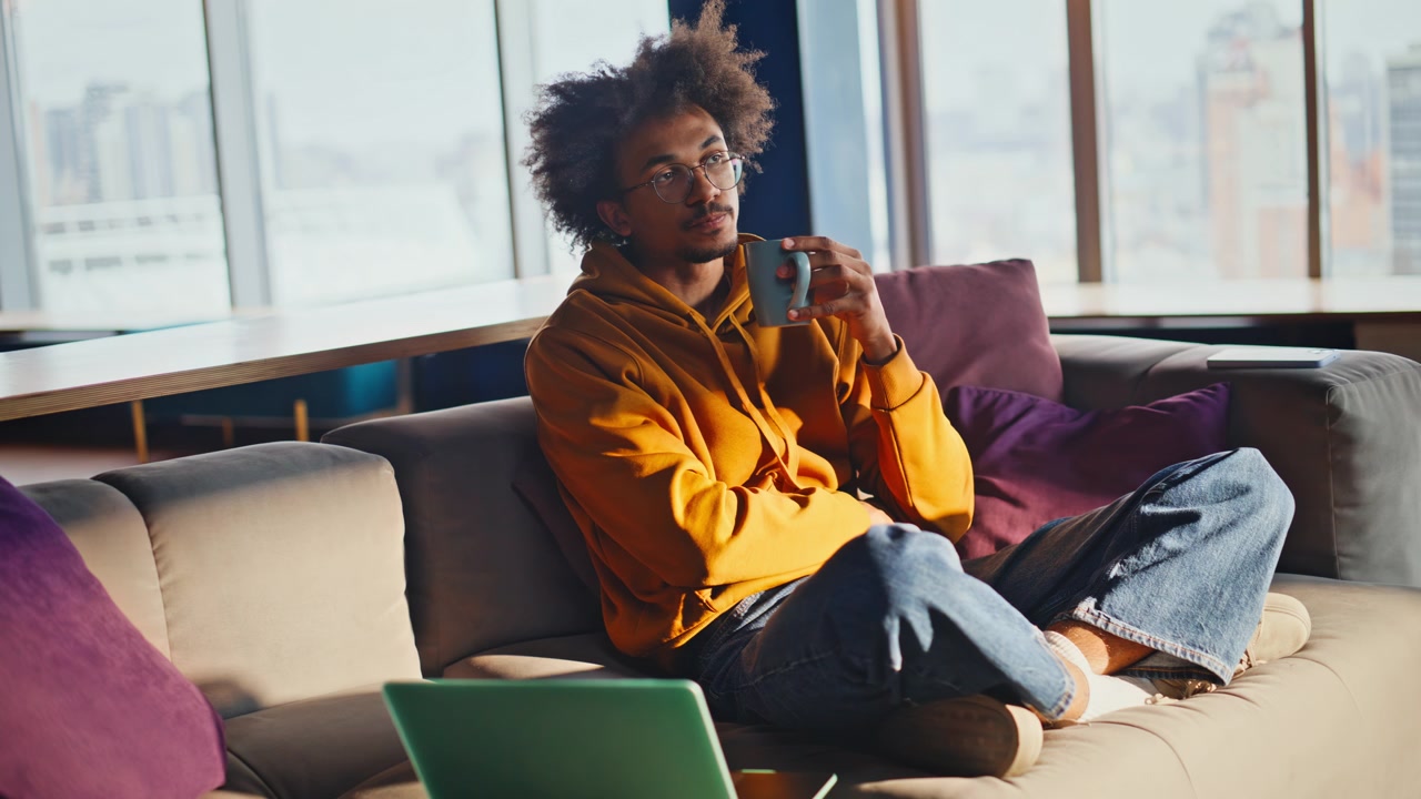 Afroamerikansk mann drikker kaffe i sofaen. Frilanser slapper av i coworking-pause