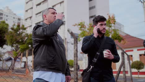 two men walking and talking outdoors while holding coffee cups, one making a phone call