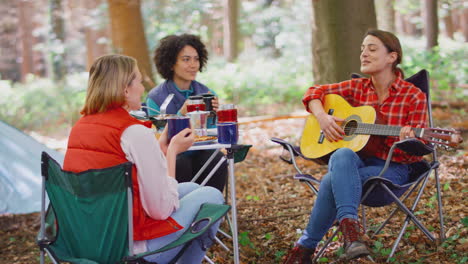 group of female friends on camping holiday in forest eating meal and singing along to guitar