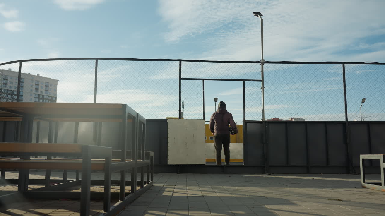 Back view of individual walking away from urban sports arena, carrying a soccer ball, with vibrant yellow storage containers and city buildings in the background, sunlight creating a glow