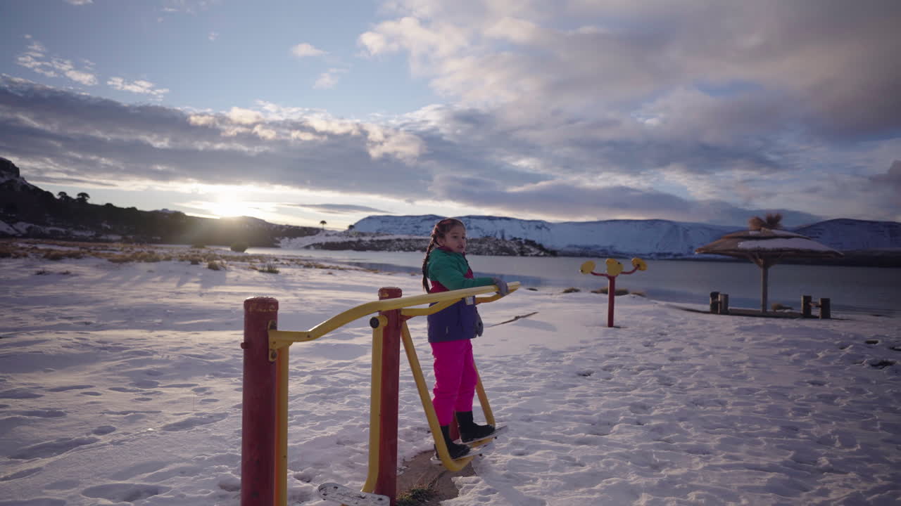 Young girl in colorful winter clothing plays on an outdoor ski simulator exercise machine beside a frozen lakeshore with mountains in Patagonia, Argentina, captured in wide ground-level shot
