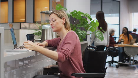 businesswoman in wheelchair working on laptop in kitchen area of busy modern office