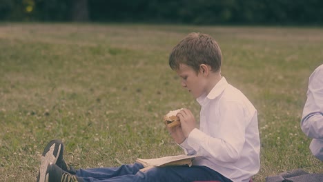 young concentrated pupil reads book eating burger on lawn