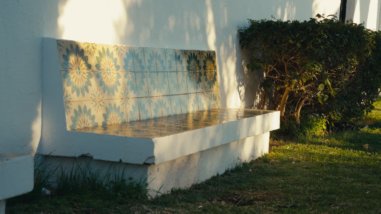Panca decorativa in ceramica con piastrelle floreali blu, immersa nella calda luce dell'ora d'oro, che proietta morbide ombre di alberi su un muro imbiancato in un tranquillo giardino spagnolo.