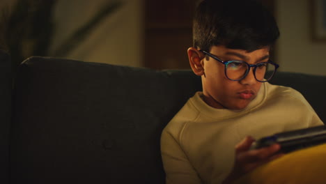 close up of young boy sitting on sofa at home playing game or streaming onto handheld gaming device at night 1