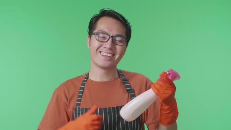 primer plano de ama de casa masculina asiática con un delantal mirando el spray en la mano luego mostrando los pulgares hacia arriba gesto a la cámara en el estudio de pantalla verde