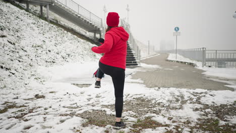 back view of woman performing energetic winter workout outdoors on snowy ground near staircase and railing, wearing red hoodie and black gloves, demonstrating fitness with a foggy background