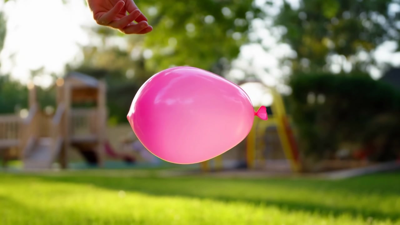 Palloncino rosa brillante tenuto da una mano in un parco all'aperto soleggiato. Il palloncino in primo piano domina l'inquadratura mentre lo sfondo rivela un prato verde, alberi e un...