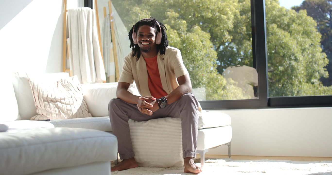 Uomo afroamericano sui vent'anni che ascolta musica con le cuffie, seduto su un pouf a casa, sorridendo.
