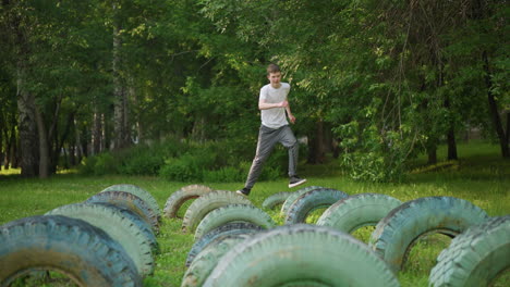 front view of a young boy in a white shirt and gray pants training on a series of tires in a grassy field, with trees and blur view of building the background